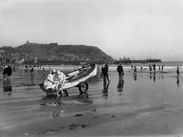 31st August 1923: Holidaymakers on the sands at Scarborough, Yorkshire. The town lies on the hill in the background. Two girls sit on a rescue boat drawn up on the sand.